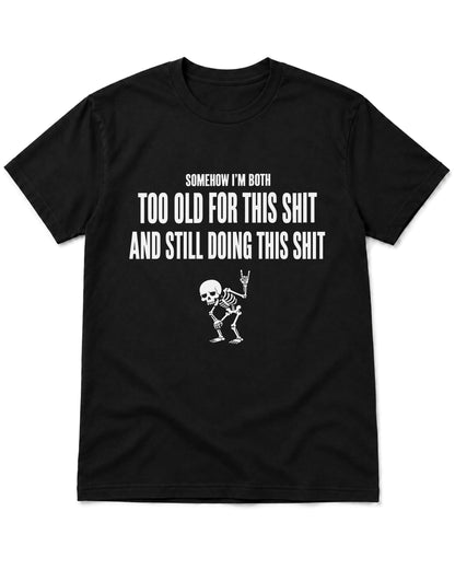 A black short-sleeve crewneck t-shirt with a white graphic print on the front. The text reads 'SOMEHOW I'M BOTH TOO OLD FOR THIS SHIT AND STILL DOING THIS SHIT' above a small white skeleton dancing and making a hand gesture.